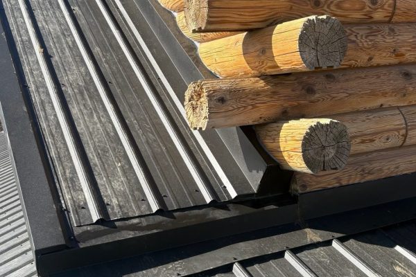 Close-up view of a standing seam metal roof transition on a log cabin, showing differing roof slopes, concealed fasteners, and black flashing where the roof meets the log walls.