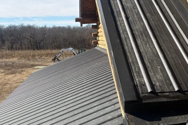 Close-up view of a standing seam metal roof transition on a log cabin, showing differing roof slopes, concealed fasteners, and black flashing where the roof meets the log walls.