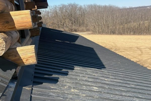 Close-up view of a standing seam metal roof transition on a log cabin, showing differing roof slopes, concealed fasteners, and black flashing where the roof meets the log walls.
