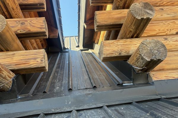 Close-up view of a standing seam metal roof transition on a log cabin, showing differing roof slopes, concealed fasteners, and black flashing where the roof meets the log walls.