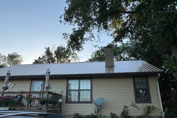 Single-family home with light beige siding, dark brown trim, a light corrugated metal roof, brick chimney, wooden deck with patio umbrellas and potted plants, and a satellite dish in a wooded setting.