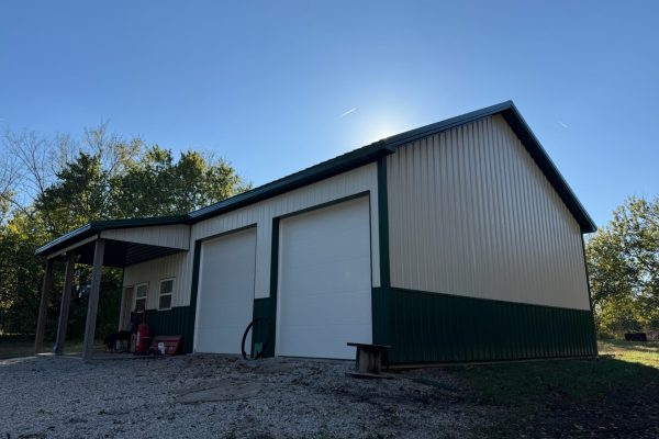 Two-tone metal pole barn or workshop with evergreen and beige vertical panels, a covered porch, two white overhead garage doors, and a walk-in entry door for durable low-maintenance use.