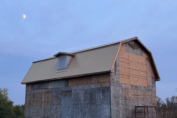 Barn with a gambrel roof and metal panels undergoing renovation, featuring weathered wood siding, new OSB sheathing, and construction materials in the foreground.