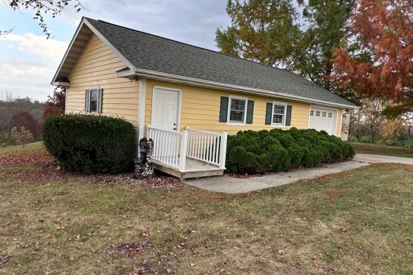 Single-story yellow house with dark shutters, dark roof, white front deck and railing, attached garage, and a spacious backyard.