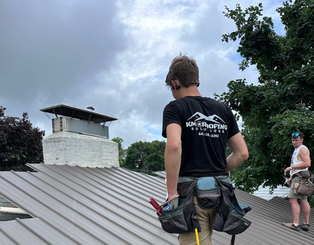 Roofers installing standing seam metal panels, including a worker wearing a tool belt and KM Roofing Solutions shirt, with a white brick chimney visible in the background.