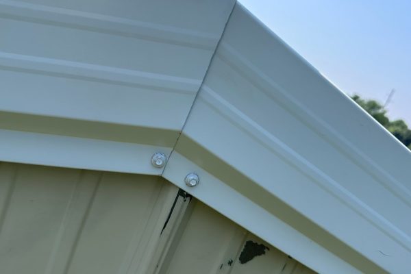 Close-up of a metal structure’s gable end and roofline with ribbed metal panels, visible flashing and trim at the eaves, and light-colored finish.