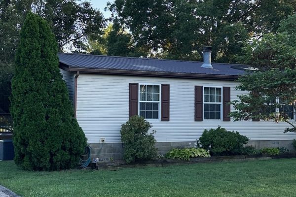 Single-story manufactured home with white horizontal siding, brown metal roof, brown shutters, front garden landscaping, and a sign near the foundation reading “Davis.”